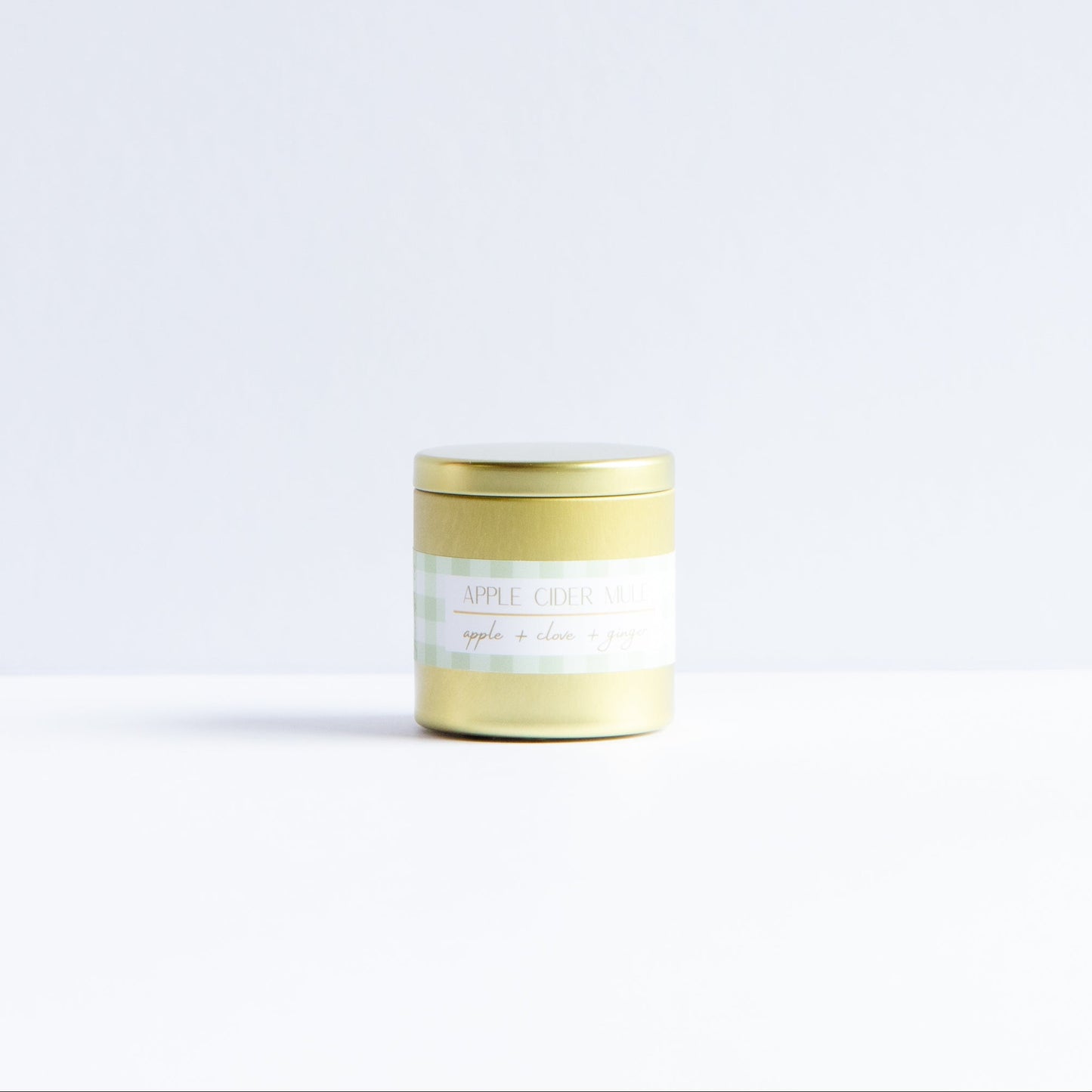A small cylindrical jar with a metallic gold lid and a white label, standing on a white surface against a very light gray background.