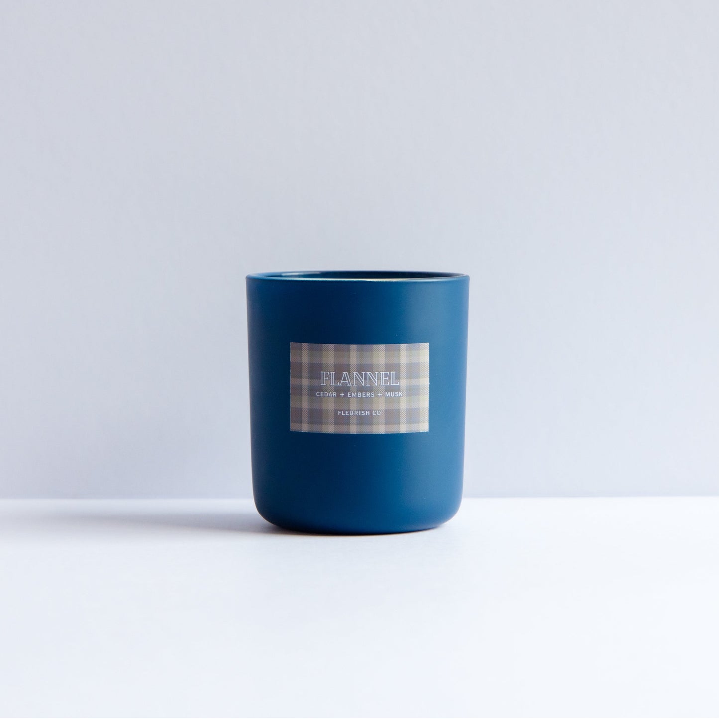 A matte dark blue glass jar candle with a white and gray plaid label reading 'DANIEL' and 'Scent of the season', set against a minimalist white and light gray background.