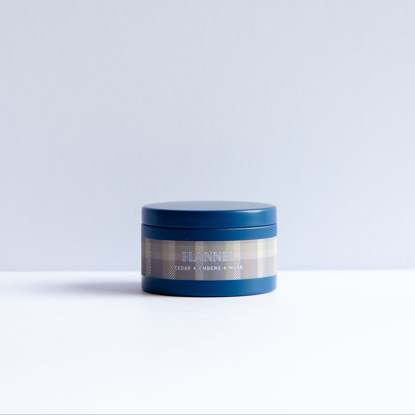 A small round blue cosmetic jar with a beige-and-blue plaid label reading 'ELANE' and 'HAIR GROWTH SCIENCE', photographed on a white surface against a pale gray background.