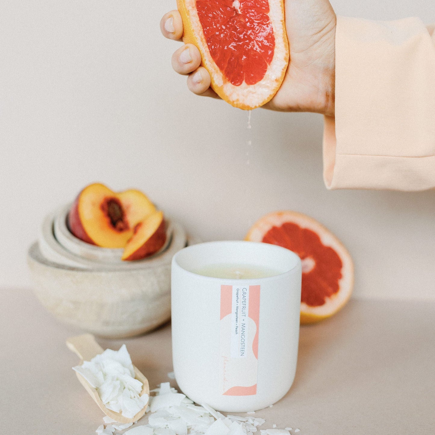 Hand holding a sliced grapefruit over a white candle with a brand label, surrounded by sea salt and peach slices on a neutral background.
