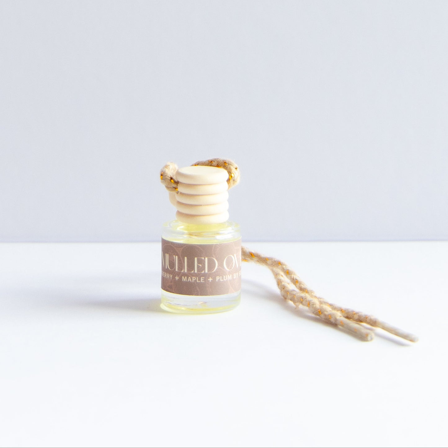 A small glass bottle of diffused oil with a tan label reading 'Mulled Over' topped with stacked natural wood beads and a braided beige cord, photographed on a white surface against a light gray background.