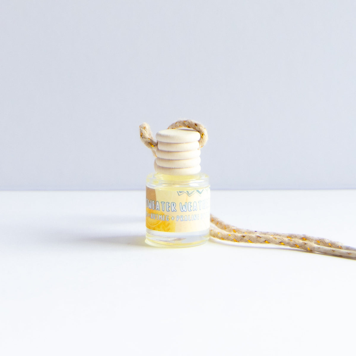 A small clear glass bottle with pale yellow liquid and a cream-colored reed diffuser stopper, shown on a white surface against a light gray background. A matching beige-and-yellow patterned cord extends to the right.