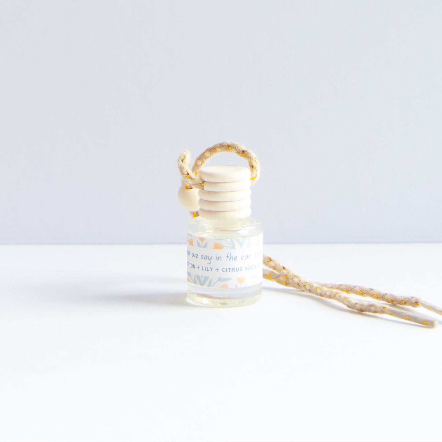 A small glass reed diffuser bottle with a light wood stopper and a matching patterned reed collar, shown on a white surface against a pale gray background. A second reed stem lies beside it.