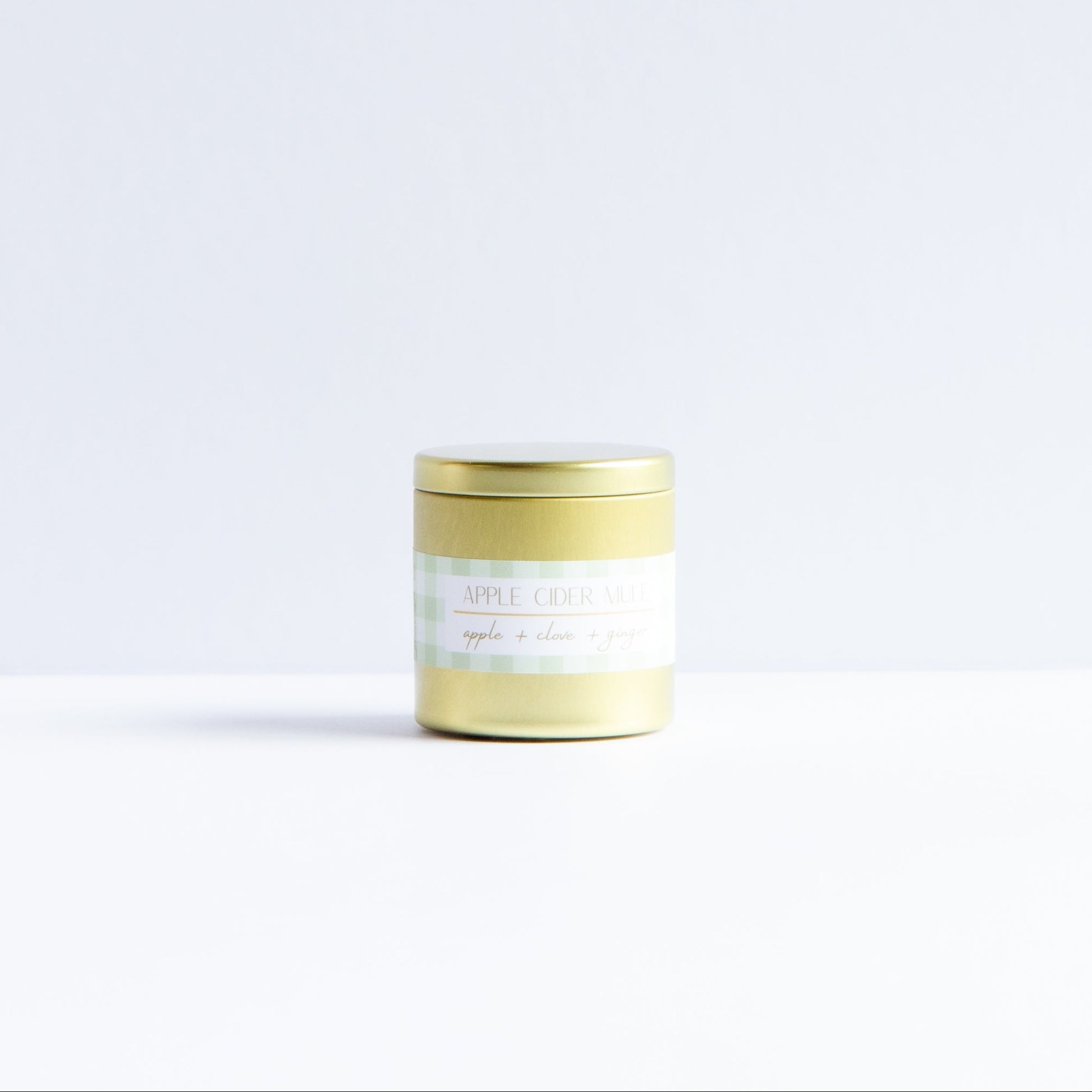 A small cylindrical jar with a metallic gold lid and a white label, standing on a white surface against a very light gray background.