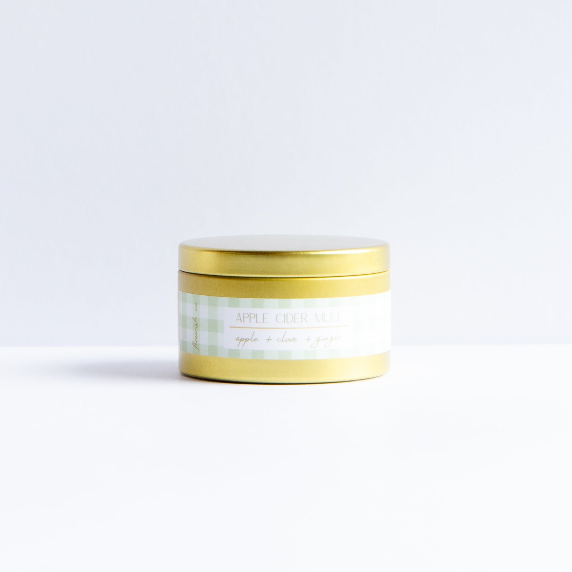 A small cylindrical cosmetic jar with a gold lid and a white label featuring a light green gingham pattern, set against a clean white background.