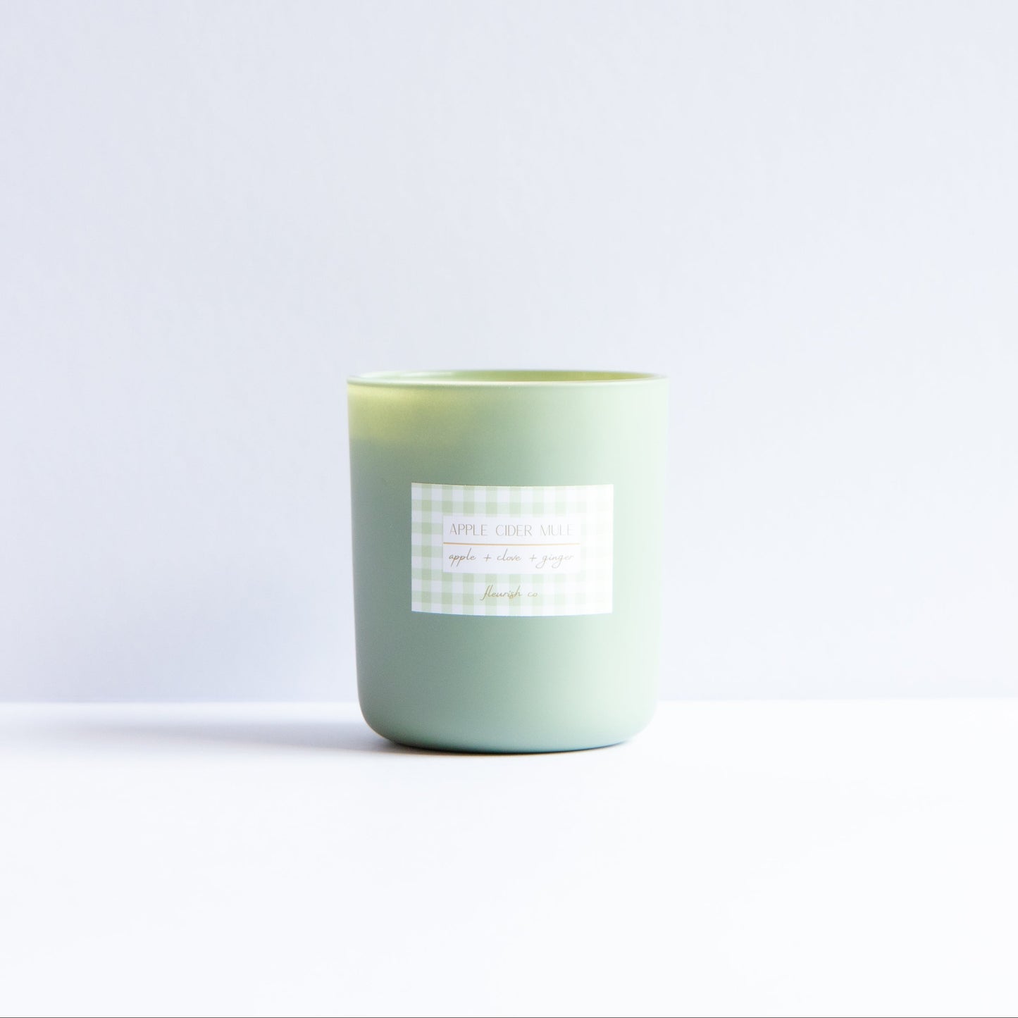 A light sage green glass jar candle with a white and gray gingham label, set against a minimalist white background.