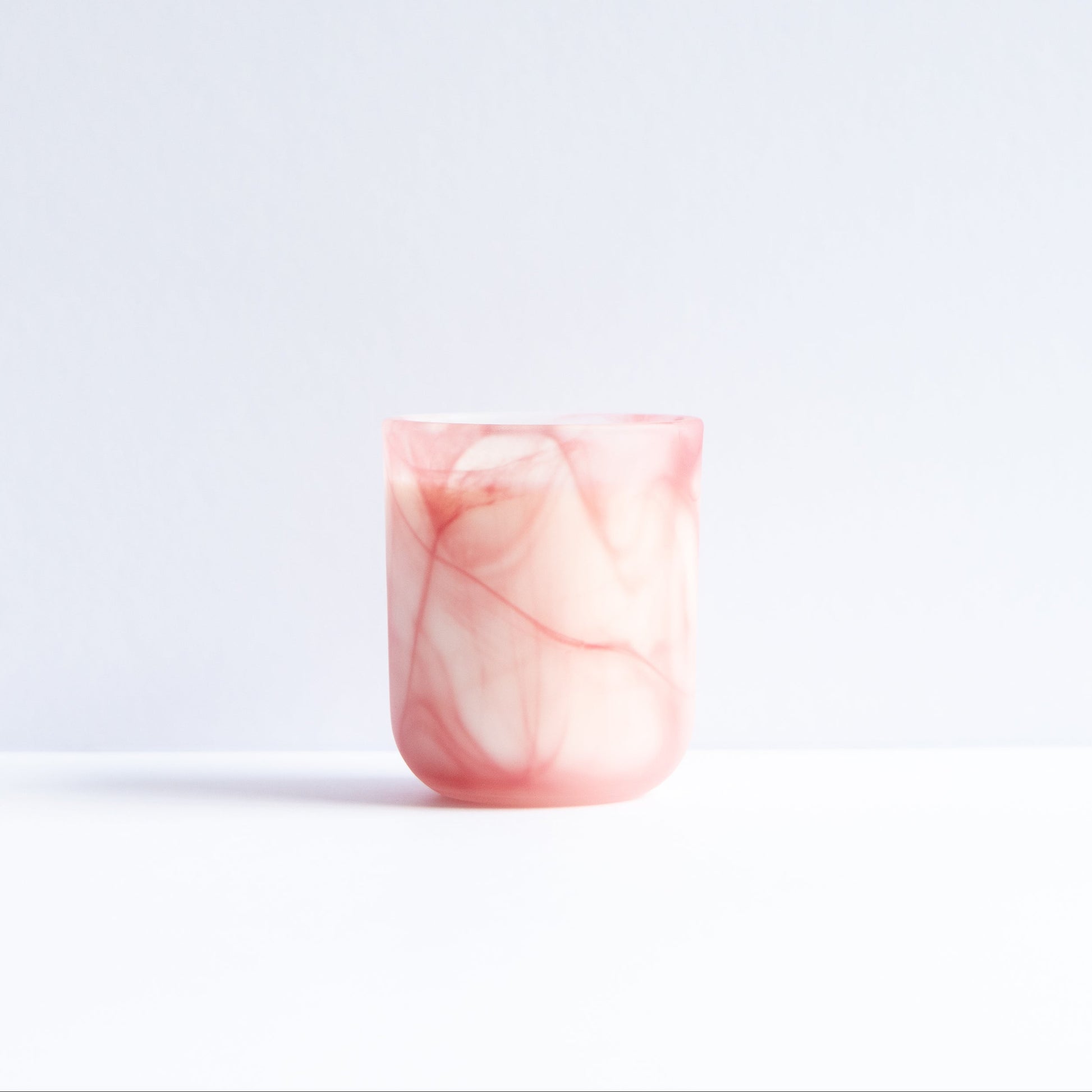 A small cylindrical cup or tumbler with a pink and white marbled pattern, standing on a white surface against a light gray background.