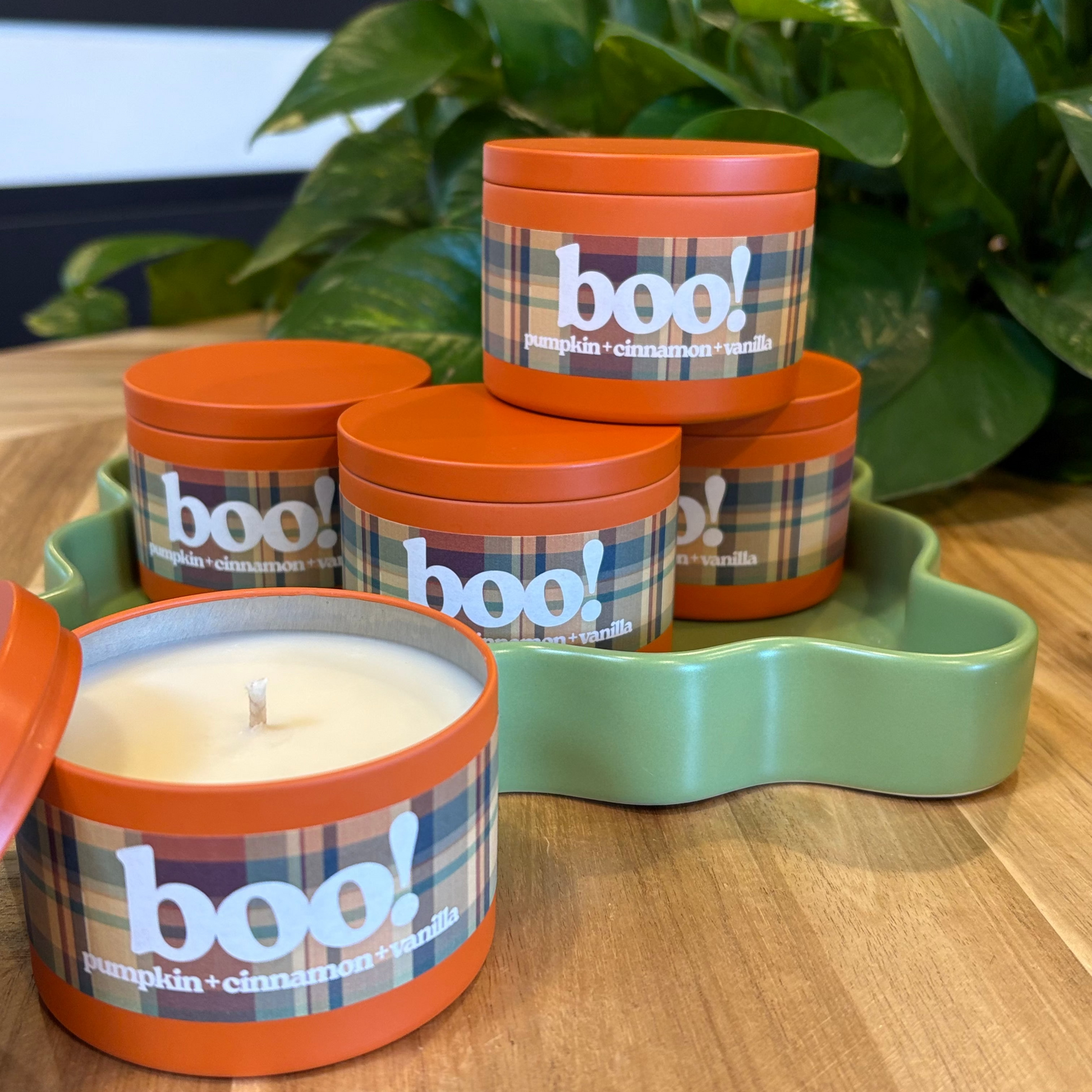 Candle in a plaid container labeled 'boo!' on a wooden surface with a plant in the background.
