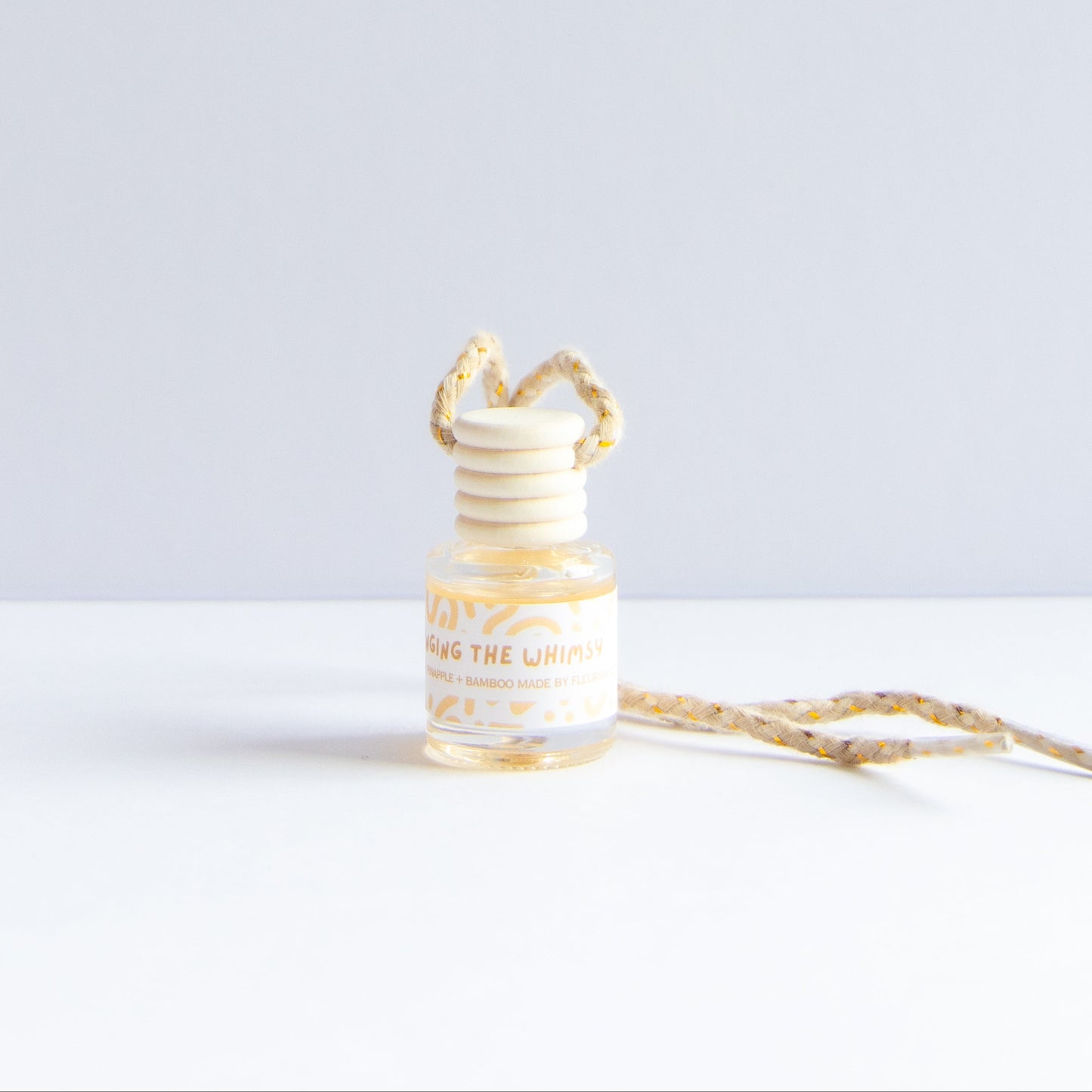 A small glass reed diffuser bottle with a cream and gold patterned reed stopper and matching ribbon, labeled 'Blossom the Whimsy', on a clean white surface against a light gray background.