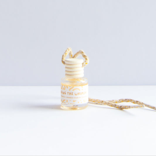 A small glass reed diffuser bottle with a cream and gold patterned reed stopper and matching ribbon, labeled 'Blossom the Whimsy', on a clean white surface against a light gray background.