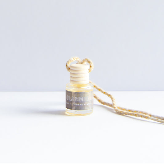 A small glass reed diffuser bottle with a light wood reed top and a braided beige-and-white hanging cord, photographed on a clean white surface against a pale gray background.
