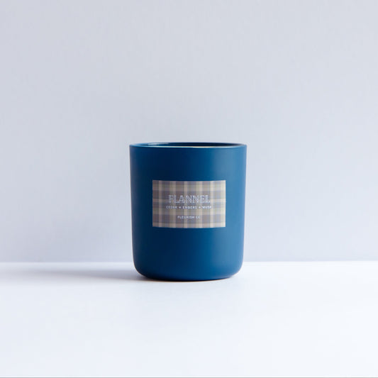 A matte dark blue glass jar candle with a white and gray plaid label reading 'DANIEL' and 'Scent of the season', set against a minimalist white and light gray background.