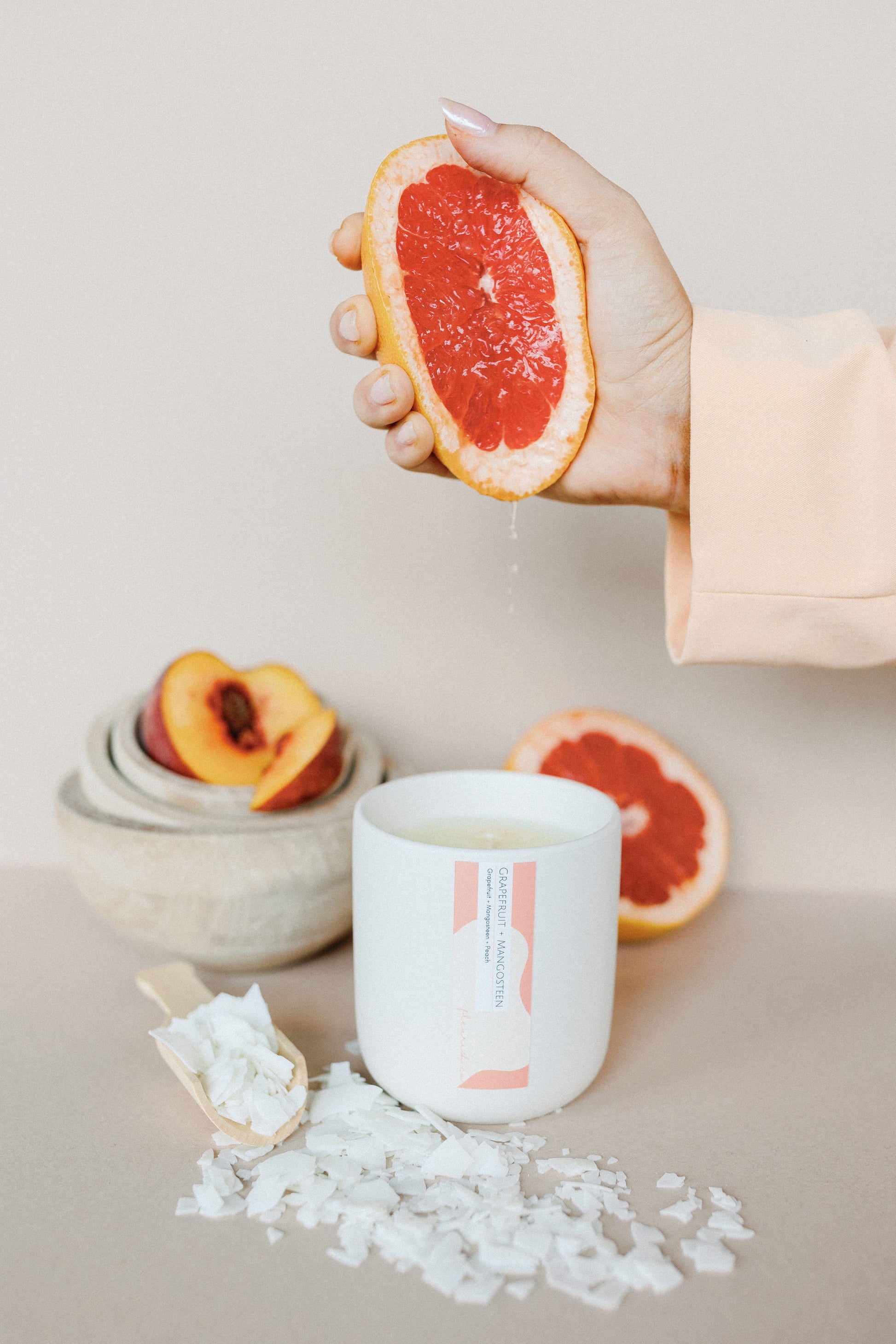 Hand holding a sliced grapefruit over a white candle with a brand label, surrounded by sea salt and peach slices on a neutral background.