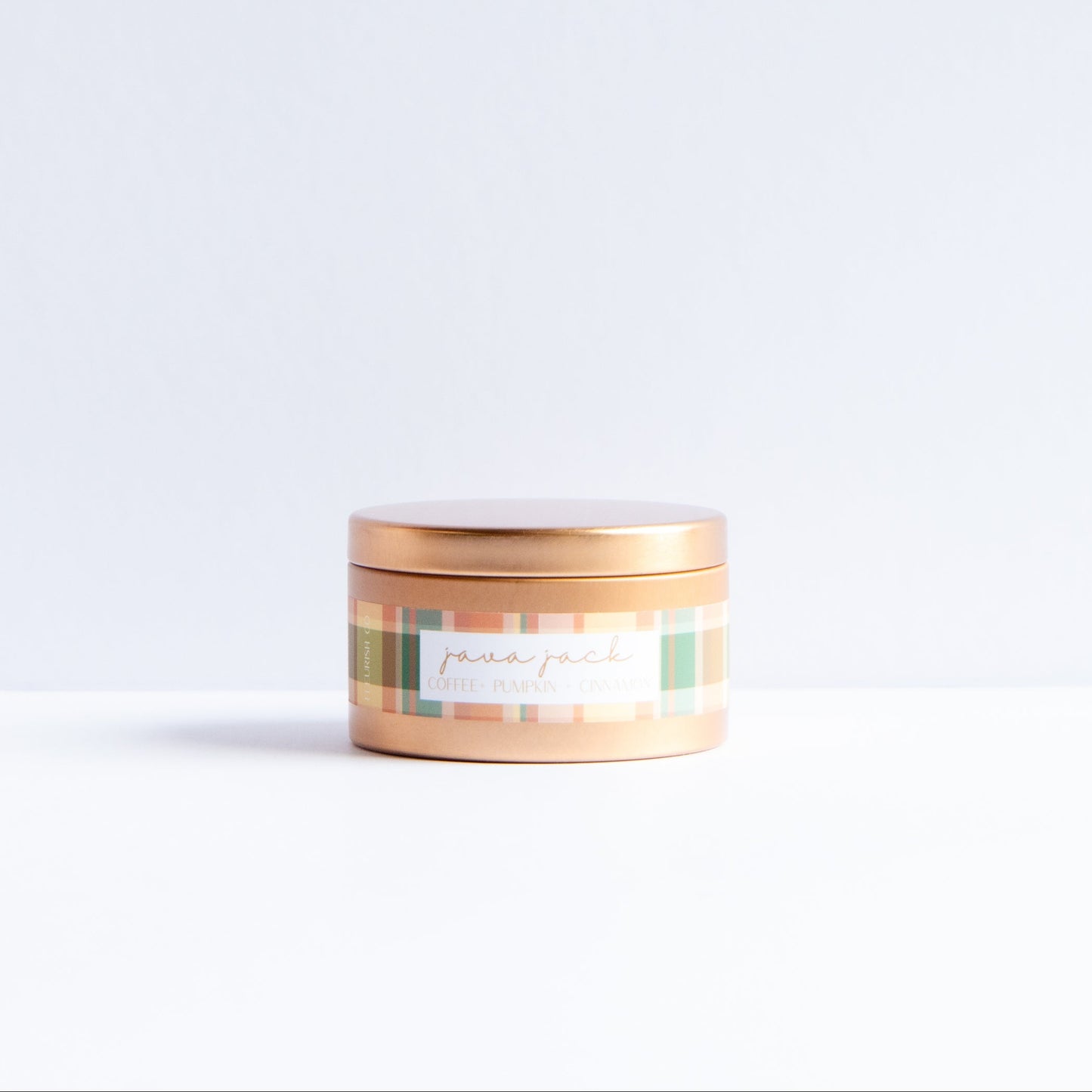 A small round tin candle with a metallic rose gold lid and a pastel plaid label reading 'Peachy Peach' on a white surface against a light gray background.