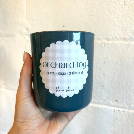 Navy blue candle with 'Orchard Fog' label held against a light tiled wall.