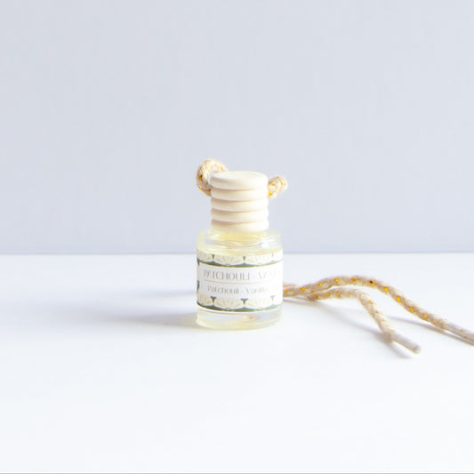A small glass reed diffuser bottle with a cream-colored wooden diffuser top and a matching cream-and-yellow patterned hanging cord, photographed on a clean white surface against a pale gray background.