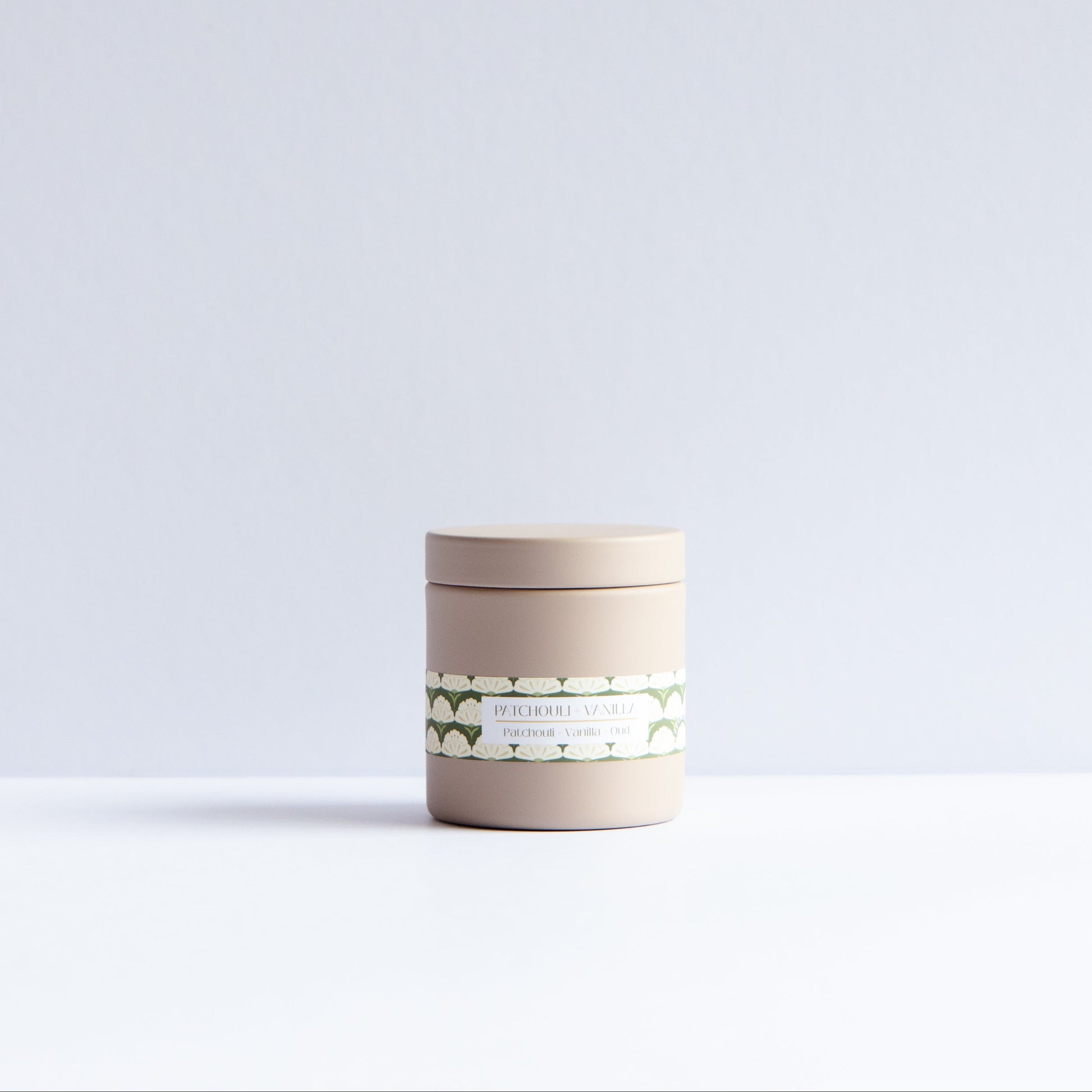 A small cylindrical beige jar with a matching lid and a decorative green and white label, centered on a clean white surface against a pale gray background.
