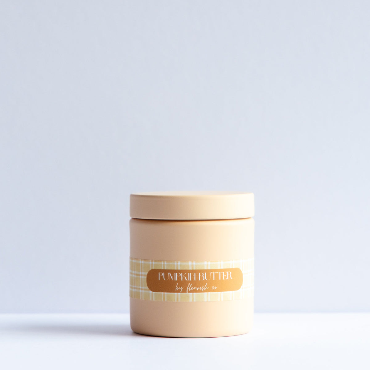 A small cylindrical jar with a matching lid, in a warm beige color, featuring a decorative label with a gold oval and a yellow plaid pattern. The label reads 'Pumpkin Spice by Emily'.