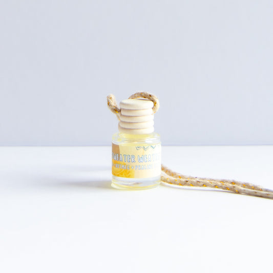 A small clear glass bottle with pale yellow liquid and a cream-colored reed diffuser stopper, shown on a white surface against a light gray background. A matching beige-and-yellow patterned cord extends to the right.