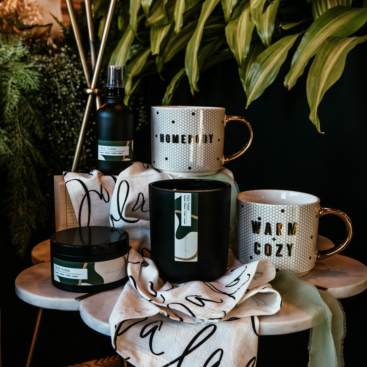 Ceramic mugs and a candle on a small table with decorative plants in the background.
