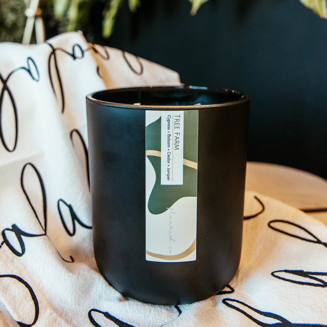 Black candle with a label on a white cloth with black text, next to a plant.