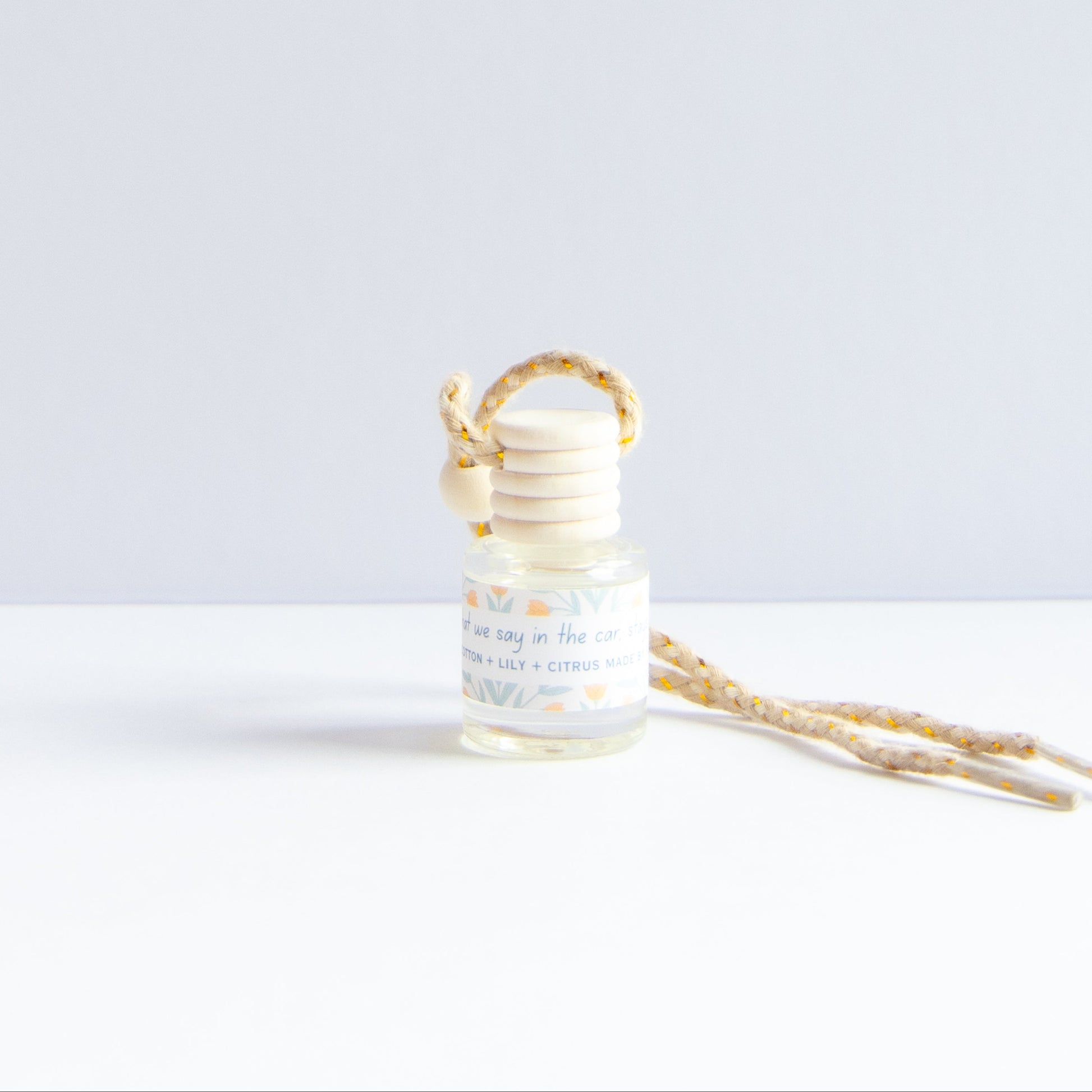 A small glass reed diffuser bottle with a light wood stopper and a matching patterned reed collar, shown on a white surface against a pale gray background. A second reed stem lies beside it.
